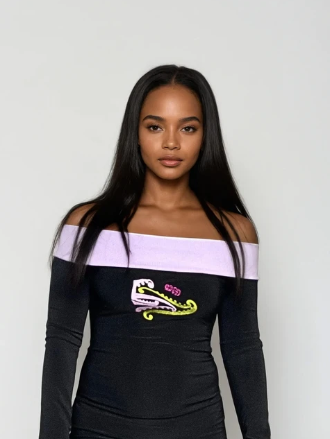 Woman wearing a black and purple dress with a ehara e te mea ko koe anake embroidery logo, standing against a white wall.